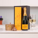Veuve Clicquot & Butlers Truffles: A bottle of Veuve Clicquot champagne is displayed alongside its signature yellow box. Two filled champagne flutes are placed beside the bottle, and a small brown candle accompanied by chocolate truffles sits in front. Veuve Clicquot & Butlers Truffles: A bottle of Veuve Clicquot champagne is displayed alongside its signature yellow box. Two filled champagne flutes are placed beside the bottle, and a small brown candle accompanied by chocolate truffles sits in front.